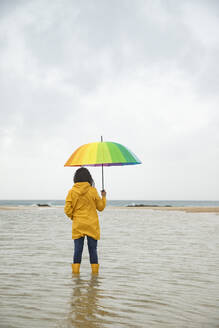 Junge Frau hält einen Regenschirm, während sie am Strand im Wasser steht - KBF00706
