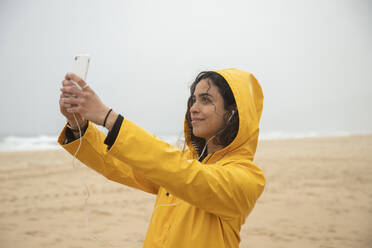 Lächelnde Frau in gelbem Regenmantel, die ein Selfie mit ihrem Mobiltelefon macht, während sie am Strand steht - KBF00708