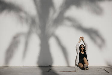 Young woman with arms raised sitting by palm tree shadow on white wall - LHPF01343