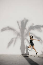 Carefree young woman jumping against palm tree shadow on white wall - LHPF01347
