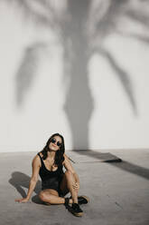 Smiling woman sitting on footpath against palm tree shadow on white wall - LHPF01348
