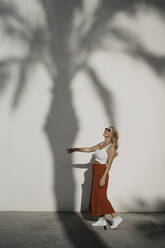 Smiling young woman walking by palm tree shadow on white wall - LHPF01351