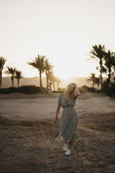 Smiling woman with hand in hair walking on beach at sunset - LHPF01362