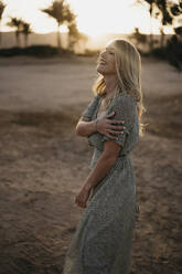 Smiling blond woman standing on beach during sunset - LHPF01364