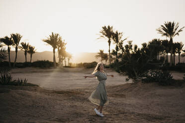 Junge Frau dreht sich mit ausgestreckten Armen am Strand bei Sonnenuntergang - LHPF01365