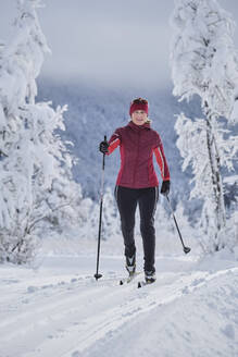 Lächelnde ältere Frau mit Skistock beim Skifahren über Schnee im Wald - MRF02475