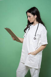 Junge Ärztin, die mit der Hand in der Tasche auf ein Klemmbrett schaut, vor grünem Hintergrund - GIOF10928
