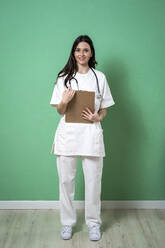 Beautiful female doctor with clipboard and stethoscope against green wall in clinic - GIOF10931