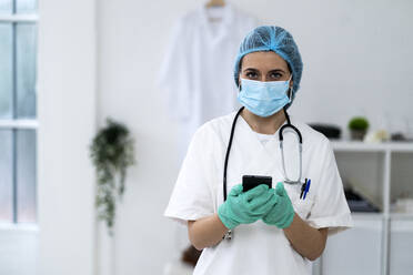 Young female doctor with surgical cap holding smart phone in clinic - GIOF10984