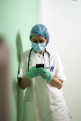 Eine Ärztin benutzt ein Smartphone, während sie sich in einer Klinik an die Wand lehnt - GIOF10988