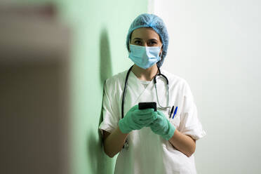 Young female doctor with mobile phone leaning by wall - GIOF10989