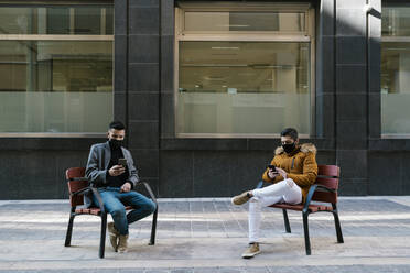 Young men wearing face mask using mobile phones while sitting on chair in city - EGAF01619
