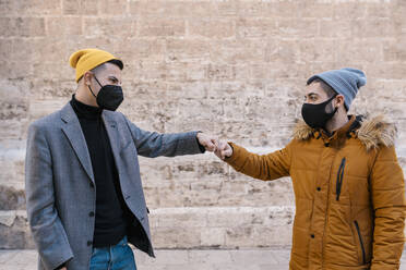 Junge Freunde mit Schutzmaske, die sich die Hände reichen, während sie an der Wand stehen - EGAF01625