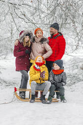 Eltern stehen bei Kindern, die im Winter im Wald auf einem Schlitten sitzen - OGF00805