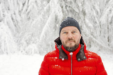 Älterer Mann mit Strickmütze, der im Wald steht und starrt, während es schneit - OGF00816