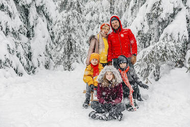 Eltern stehen bei Kindern, die im Schnee im Wald sitzen - OGF00820