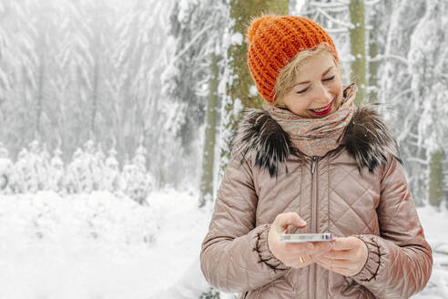 Smiling woman wearing knit hat holding mobile phone while standing in forest during winter - OGF00821