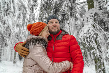 Lächelnde Frau, die einen Mann umarmt, während sie im Winter im Wald steht - OGF00824