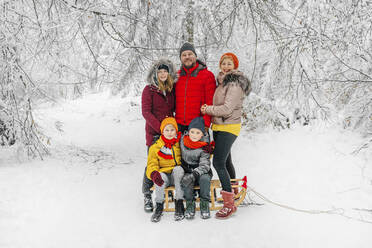 Lächelnde Eltern stehen bei Kindern, die im Winter im Wald auf dem Schlitten sitzen - OGF00825