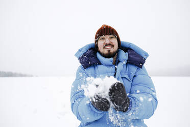 Lächelnder junger Mann mit wattierter Jacke beim Spielen im Schnee - KMKF01509