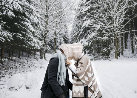 Romantisches Paar, das sein Gesicht mit einem Schal bedeckt, während es auf dem Schnee im Wald steht - KMKF01521