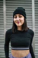 Smiling young woman with knit hat standing against metallic shutter - GRCF00679