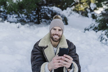 Smiling young man holding mobile phone while sitting in snow - EBBF02382