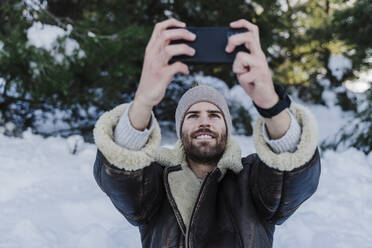 Lächelnder Mann in warmer Kleidung, der im Winter ein Selfie mit seinem Smartphone macht - EBBF02383