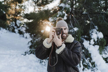 Mann in warmer Jacke, der durch die Kamera fotografiert, während er gegen Bäume im Schnee steht - EBBF02385