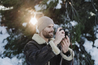Handsome man photographing through camera against trees during winter - EBBF02390
