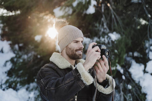 Hübscher Mann fotografiert durch die Kamera gegen Bäume im Winter - EBBF02390