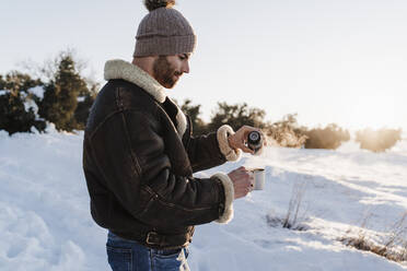 Junger Mann mit Jacke gießt Kaffee aus einer Thermoskanne in eine Tasse, während er im Schnee steht - EBBF02391