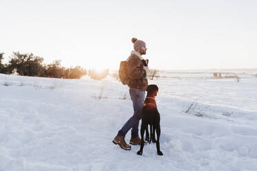 Male backpacker standing with Great Dane dog while looking away in snow - EBBF02393