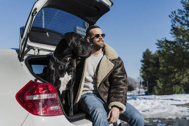 Lächelnder Mann mit Dogge, der im Kofferraum eines Autos sitzt und wegschaut, an einem sonnigen Tag - EBBF02399