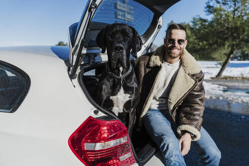 Junger Mann mit Hund sitzt im Kofferraum eines Autos an einem sonnigen Tag - EBBF02400