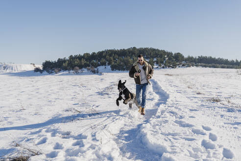 Verspielter Mann mit Hund läuft im Schnee an einem sonnigen Tag - EBBF02401