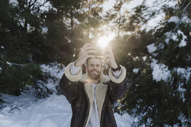 Man wearing jacket photographing while standing against trees during winter - EBBF02404