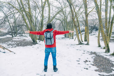 Male hiker with arms outstretched carrying backpack standing in snowy forest - IFRF00358