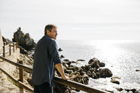 Mature man leaning on railing while looking at sea during sunny day - JRFF05004