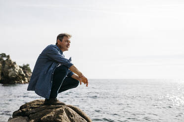 Man crouching on rock against clear sky during sunny day - JRFF05011