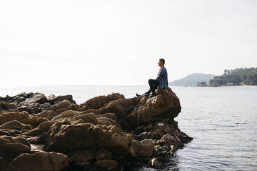 Mature man sitting on rock against clear sky - JRFF05014