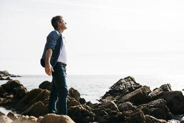 Enjoying mature man standing amidst rock by sea during sunny day - JRFF05015