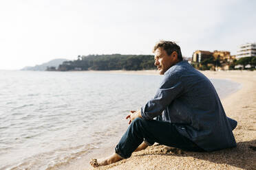 Mann sitzt am Strand auf Sand am Rande des Wassers an einem sonnigen Tag - JRFF05023