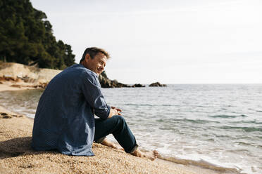 Smiling man sitting on beach while looking away against sea at beach - JRFF05024
