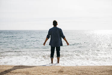 Man with arms outstretched standing at water's edge against clear sky - JRFF05028