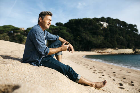 Lächelnder Mann mit Smartphone sitzt am Strand gegen Hügel während sonnigen Tag - JRFF05033