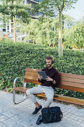 Geschäftsmann mit Kopfhörern, der ein digitales Tablet benutzt, während er auf einer Bank sitzt - BOYF01648