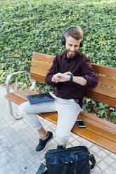 Mid adult businessman wearing headphones checking time while sitting with digital tablet on bench - BOYF01649
