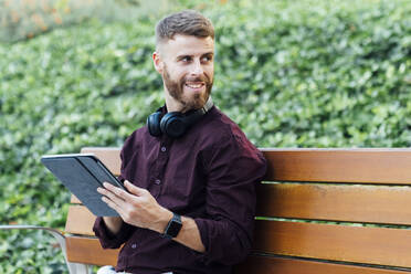 Geschäftsmann mit digitalem Tablet und Kopfhörern, der wegschaut, während er im Park sitzt - BOYF01651