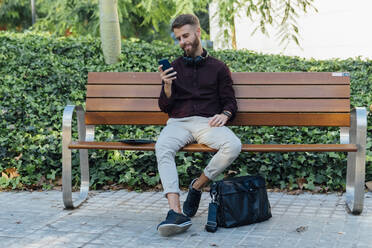 Geschäftsmann, der ein Mobiltelefon benutzt, während er auf einer Bank im Park sitzt - BOYF01652